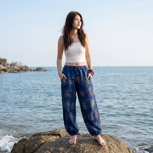 Female model wearing blue stonewashed organic cotton harem pants, posing on coastal rocks at a Thailand beach.