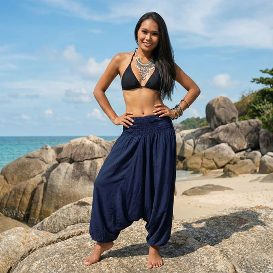 Woman in black bikini top and blue thai jumpsuit harem pants standing on a rocky beach with ocean and sky in the background