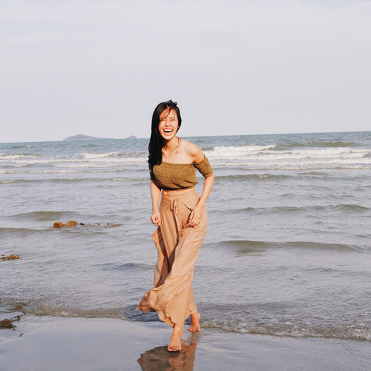 Woman in beige Thai wrap pants skirt with brown top standing in the ocean with a light sky above.