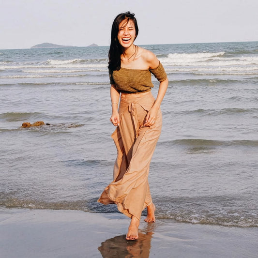Woman in beige Thai wrap pants skirt with brown top standing in the ocean with a light sky above.