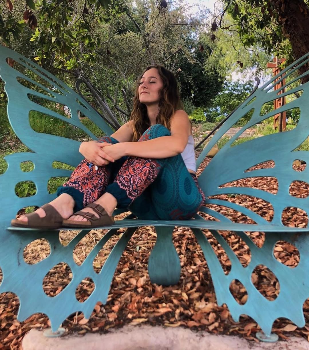 Woman wearing turquoise Thai harem yoga pants with mandala print and grey top sitting on a butterfly-shaped metal bench in a park