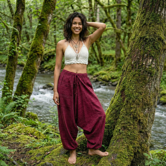 Woman standing in a forest by a stream wearing a white top and maroon pinstripe thai harem pants.