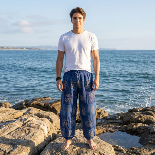 Male model wearing blue stonewashed organic cotton harem pants, posing on coastal rocks at a Thailand beach.