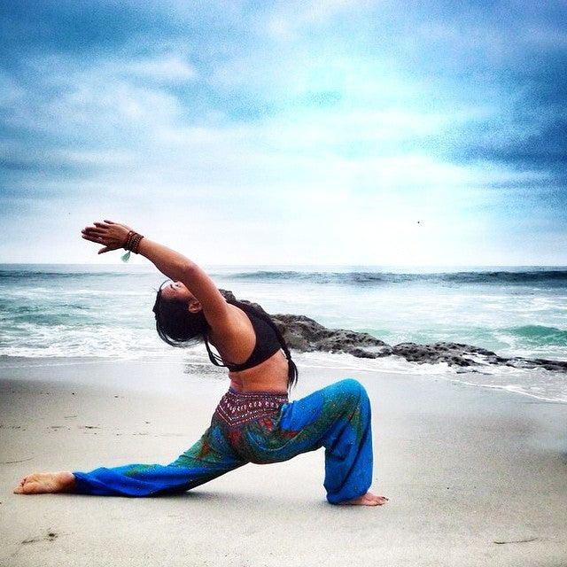 Person practicing yoga on a beach with ocean and sky in the background, wearing a blue Thai harem yoga pants with peacock print and  black top