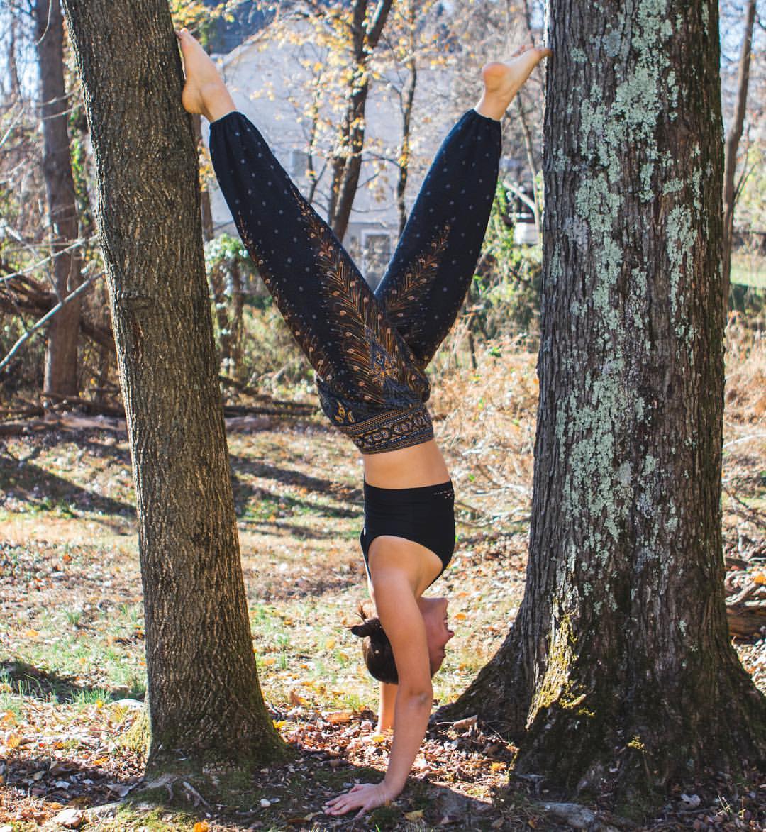 Woman green Thai harem yoga pants with peacock print and black top doing a handstand between two trees in a natural setting