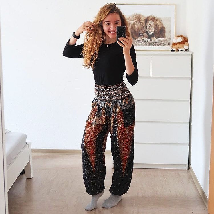 Woman taking a mirror selfie wearing a green Thai harem yoga pants with peacock print and black top in a room.