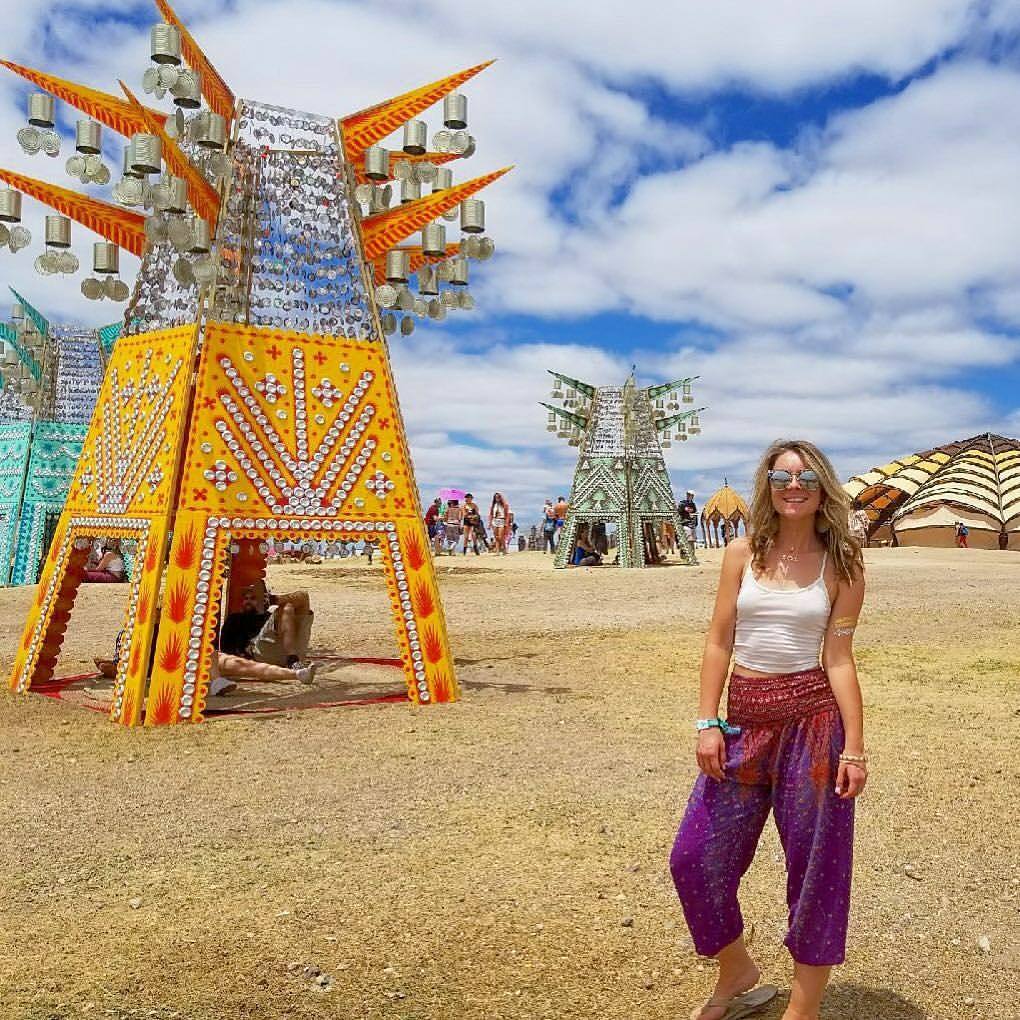 Woman in purple Thai harem yoga pants with peacock print and white top posing in front of a colorful art installation at a festival.