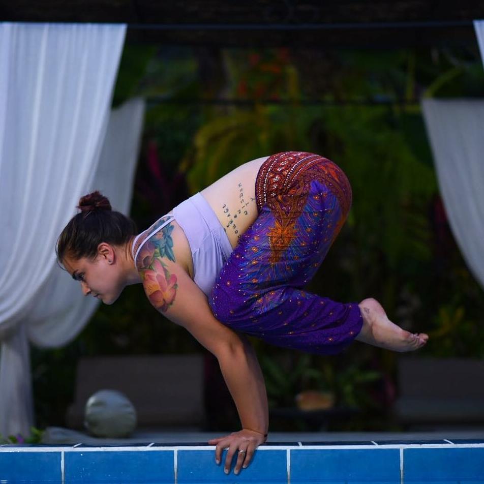 Woman in a yoga pose by a pool with a blurred background wearing purple Thai harem yoga pants with peacock print and white top