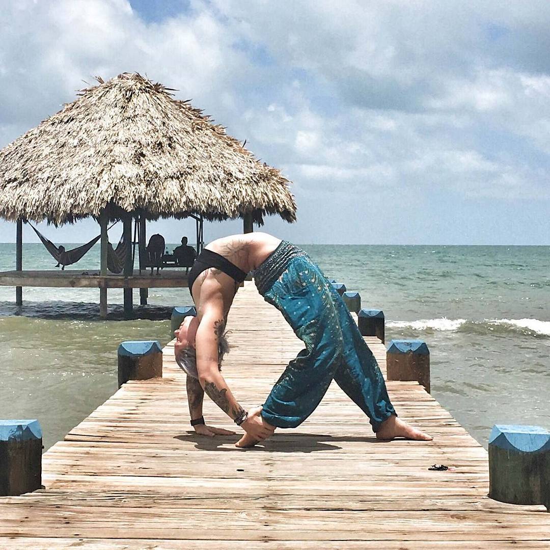 Woman in turquoise Thai harem yoga pants with peacock print and black top doing a handstand on a wooden dock with a thatched umbrella and hammock in the background.