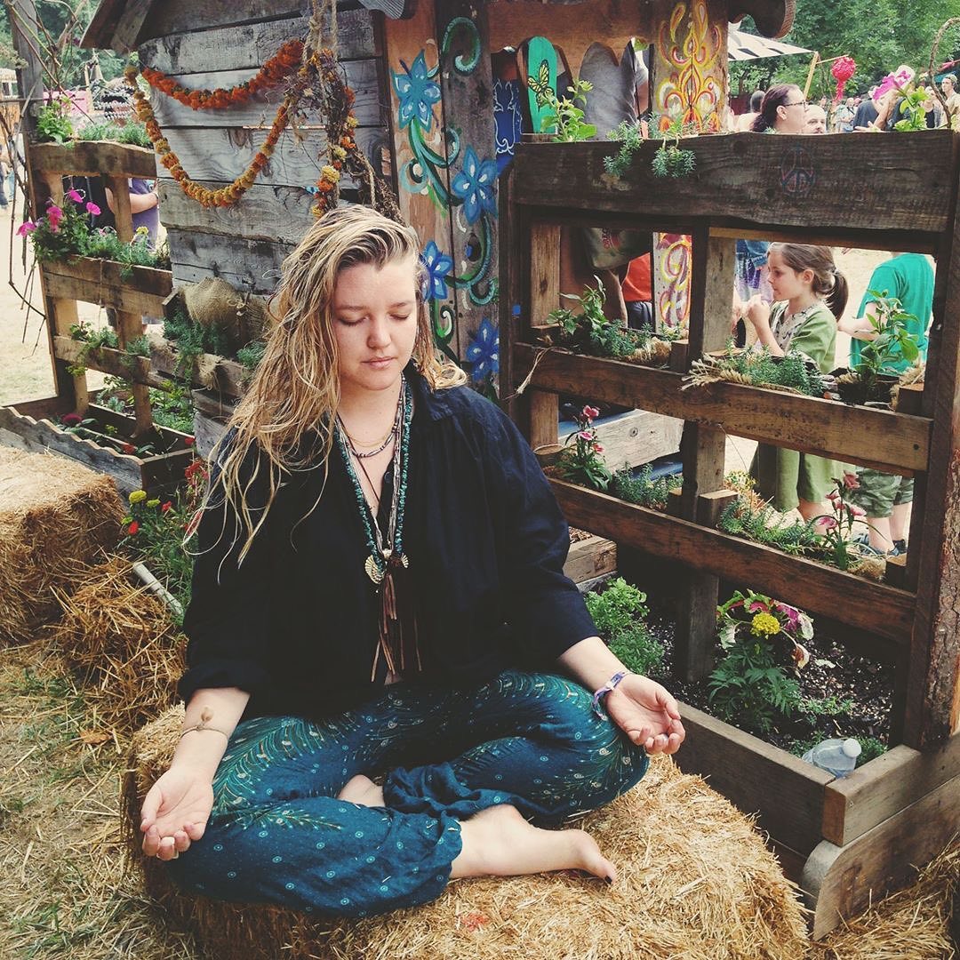 Woman turquoise Thai harem yoga pants with peacock print and black jacket meditating on a hay bale in a rustic outdoor setting with wooden structures and plants.