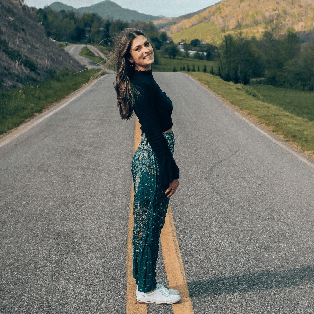 Woman turquoise Thai harem yoga pants with peacock print and black top standing on a road with a scenic background