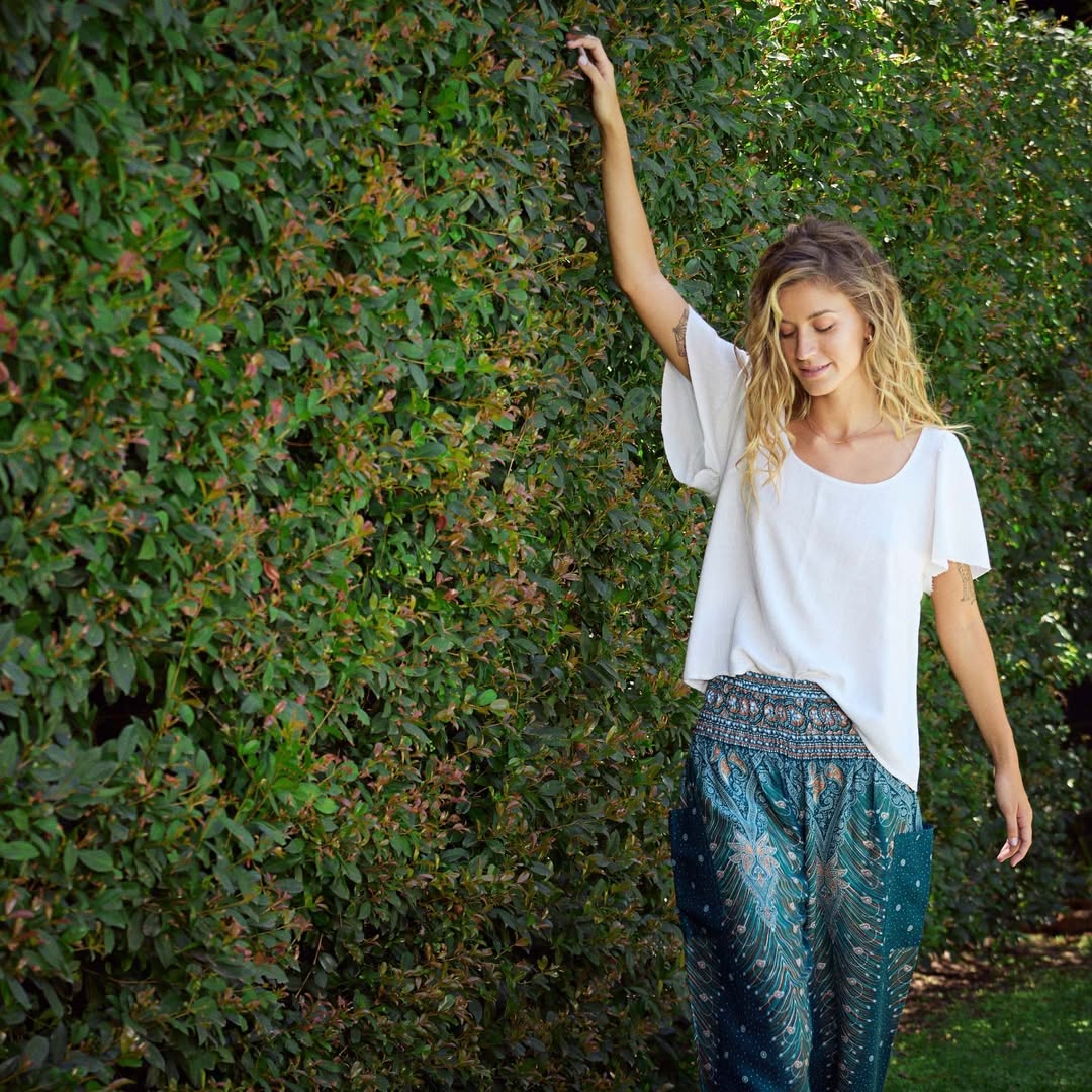 Woman in a white top and turquoise Thai harem yoga pants with peacock print standing against a green hedge