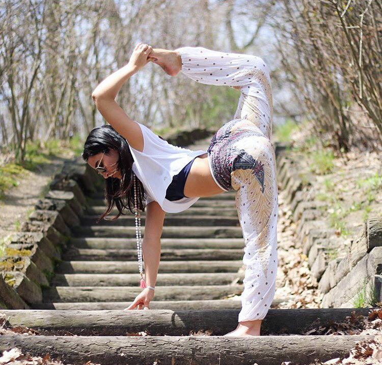 Woman in white Thai harem yoga pants with peacock print and white top practicing yoga outdoors on a wooden log with trees in the background