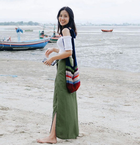 Woman in green Thai wrap pants skirt and white top standing on a beach with ocean waves and a boats in the background