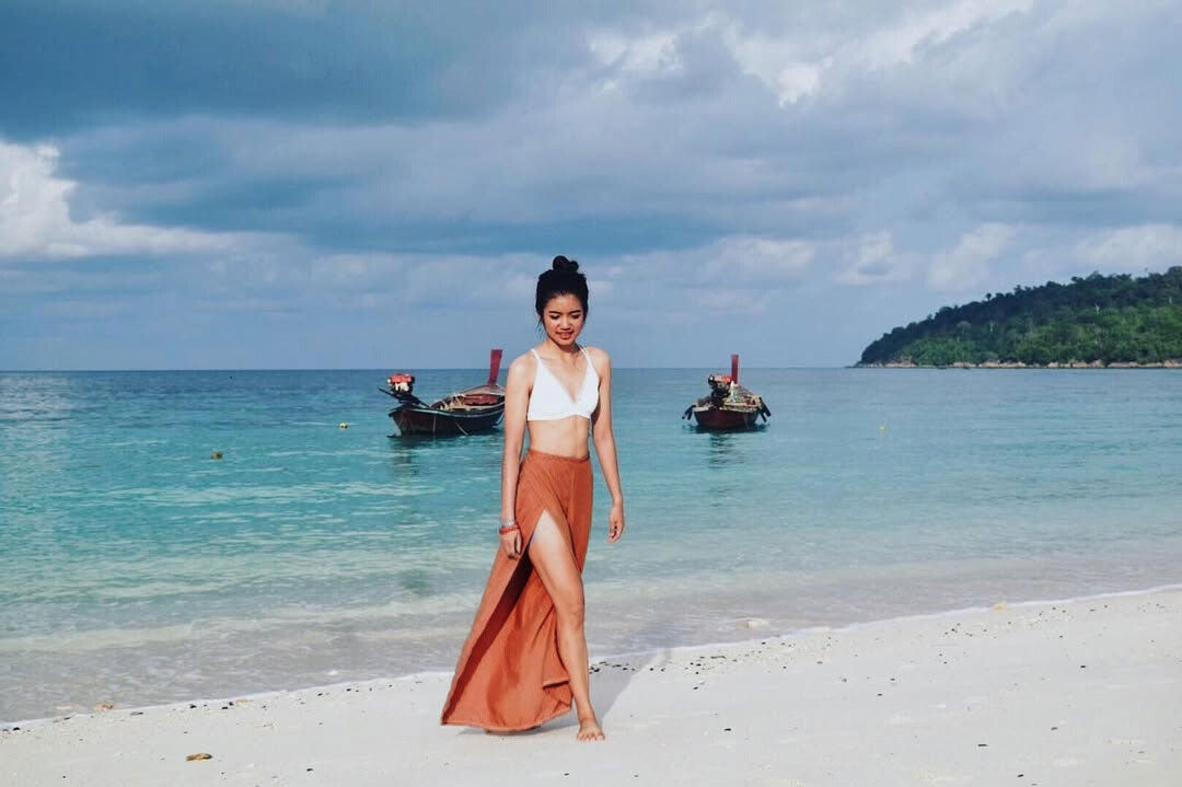 Woman in a white top and orange thai wrap pants skirt standing on a beach with boats and ocean in the background