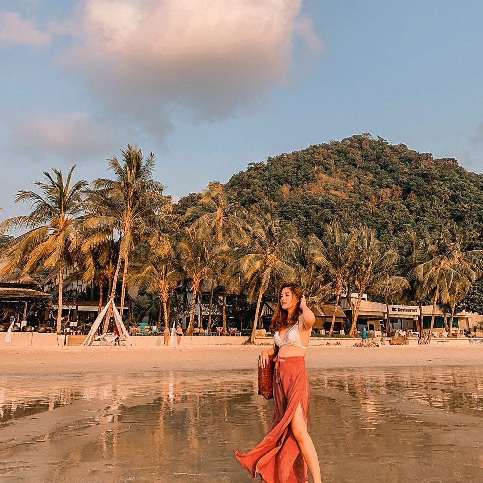 Woman in orange Thai wrap pants skirt and white top standing on a beach with palm trees and mountains in the background