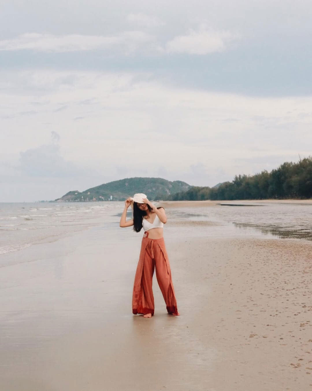 Woman in orange Thai wrap pants skirt and white top standing standing on a beach with mountains in the background