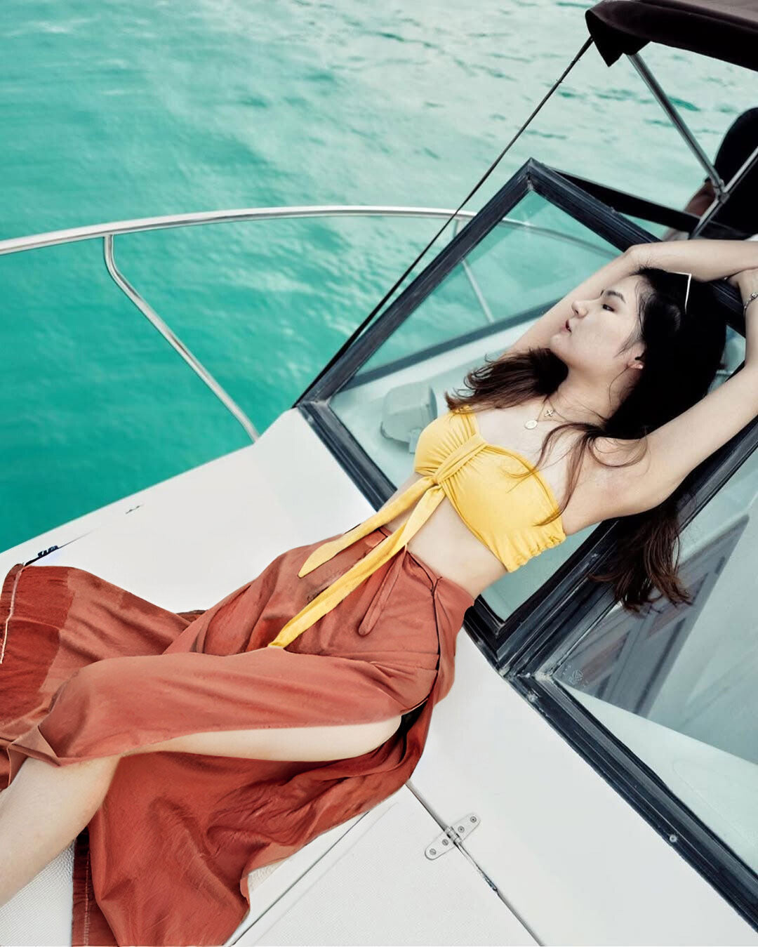 Woman in a yellow top and orange thai wrap pants skirt lying on a boat with turquoise water in the background