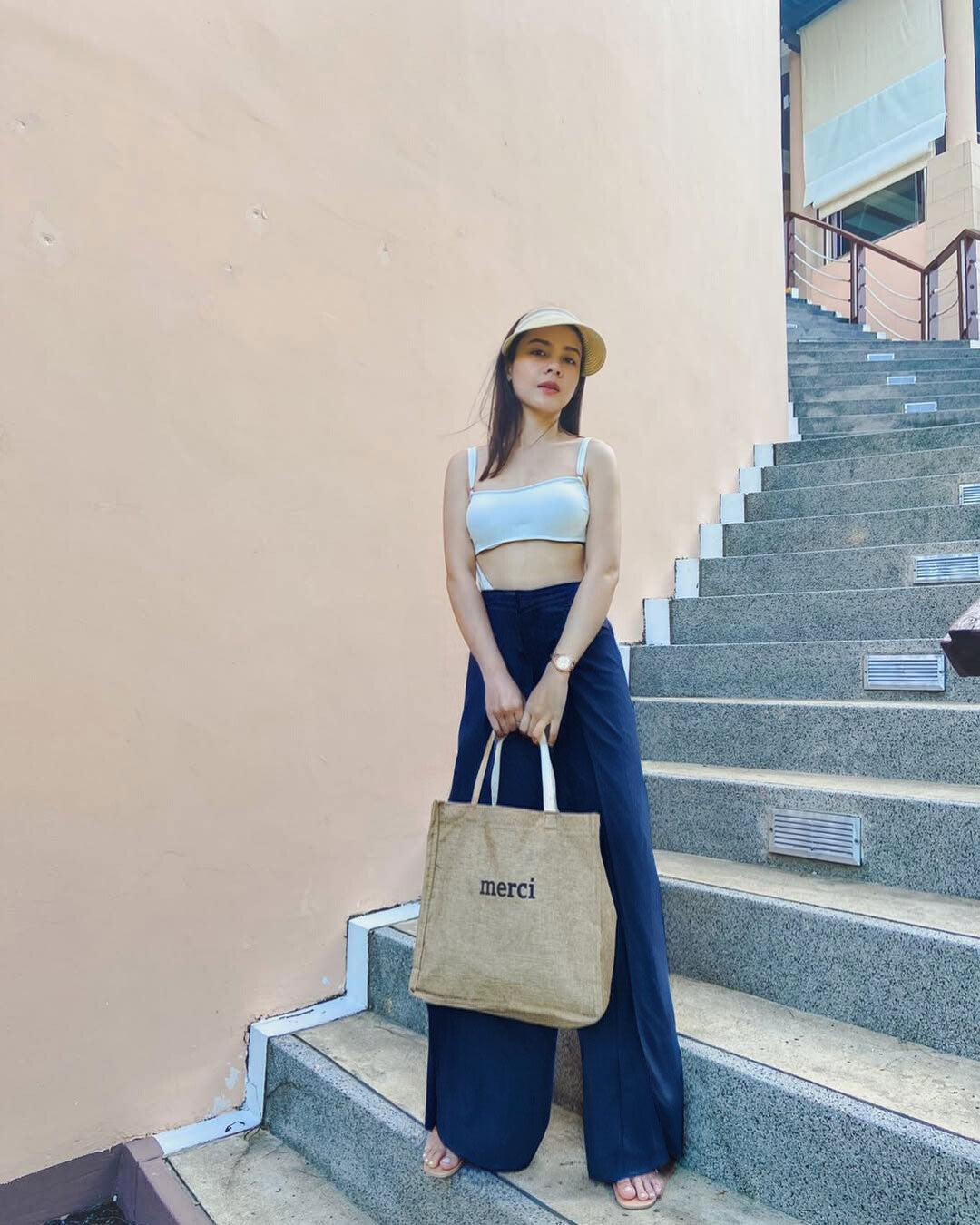 Woman in blue Thai wrap pants skirt and white top holding a 'merci' bag on a staircase