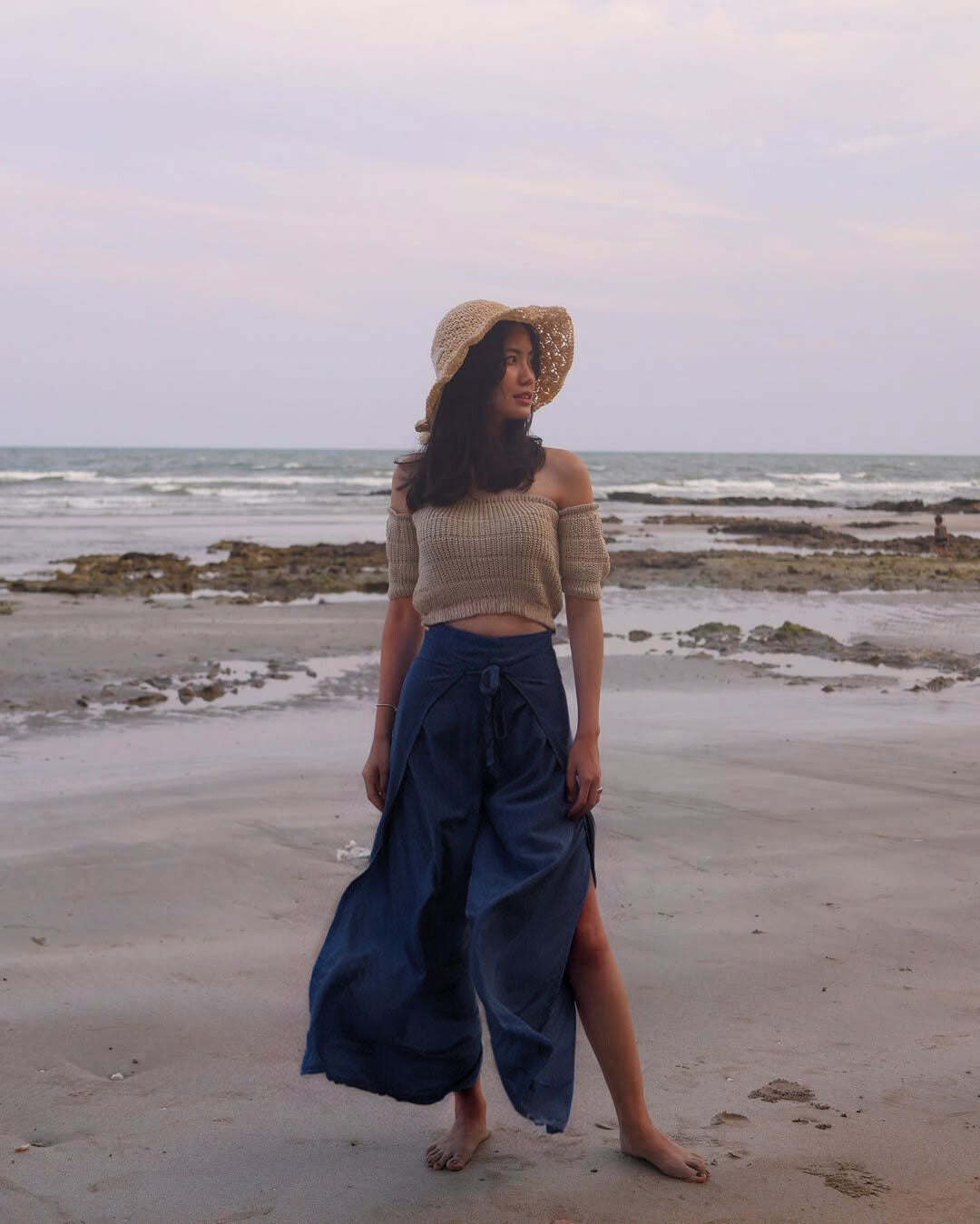 Woman standing on a beach wearing a hat and  blue Thai wrap pants skirt and brown top