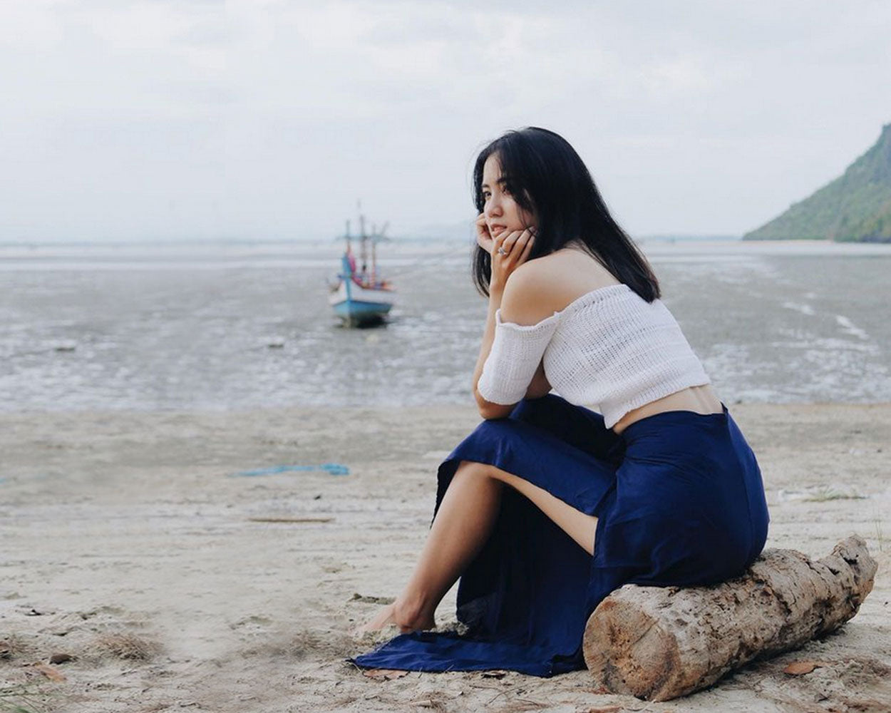 Woman in blue Thai wrap pants skirt and white top sitting on a log by the water with a boat in the background