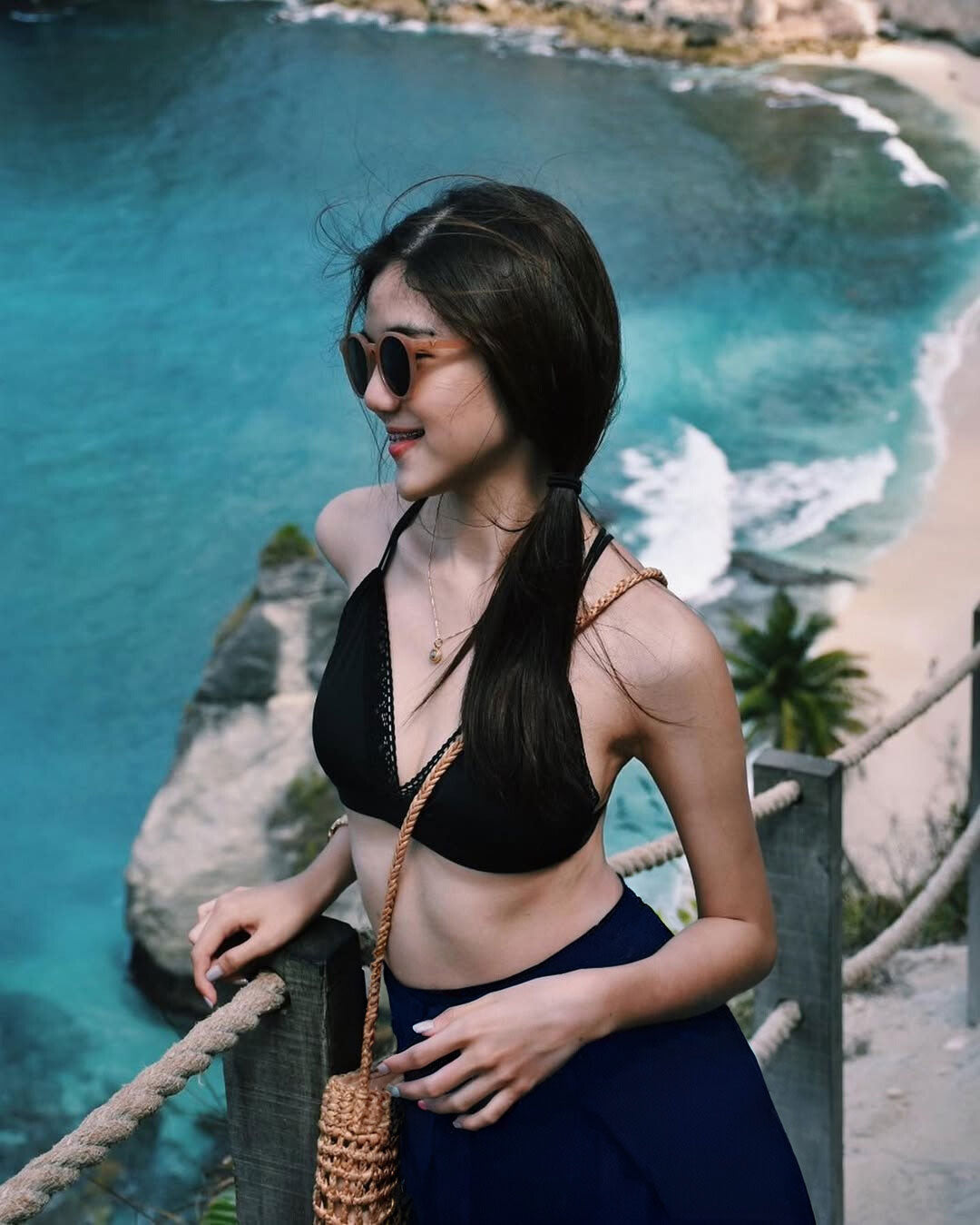 Woman in a black bikini top and blue Thai wrap pants skirt standing by a scenic beach with turquoise water and palm trees.