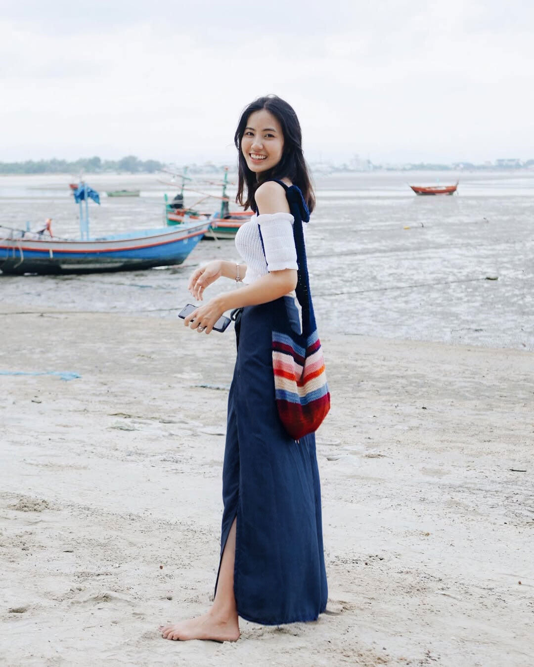 Woman in blue Thai wrap pants skirt and white top standing on a beach holding a colorful bag, with boats in the background.