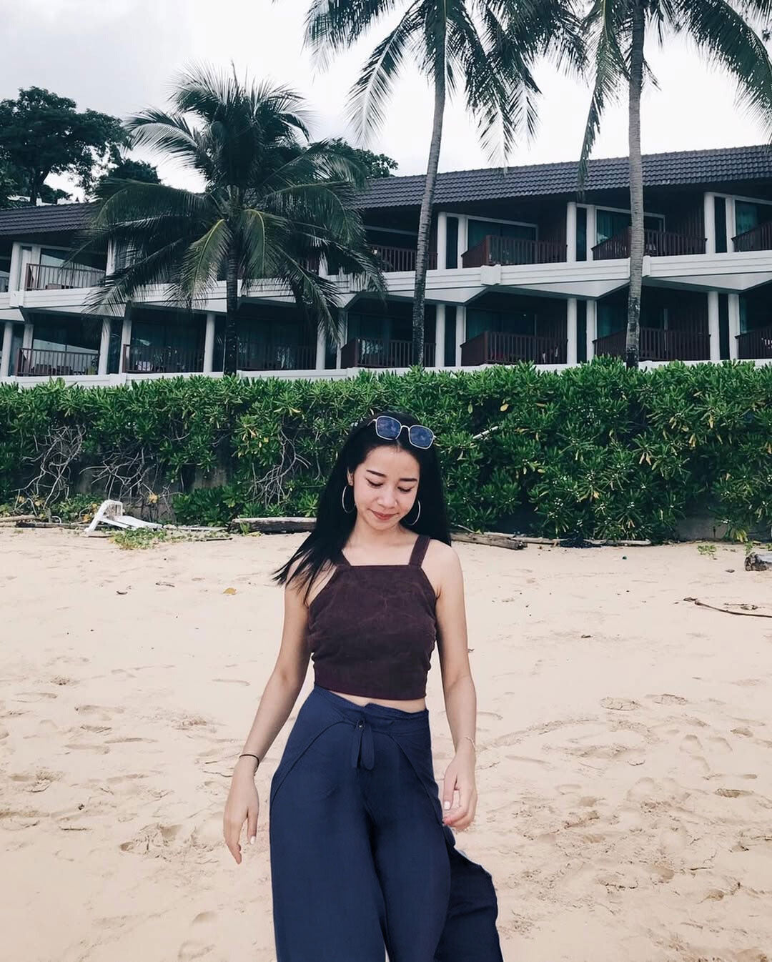Woman in blue Thai wrap pants skirt and brown top standing on a beach with palm trees and a building in the background