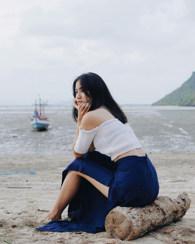 Woman in blue Thai wrap pants skirt and white top sitting on a log by the water with a boat in the background