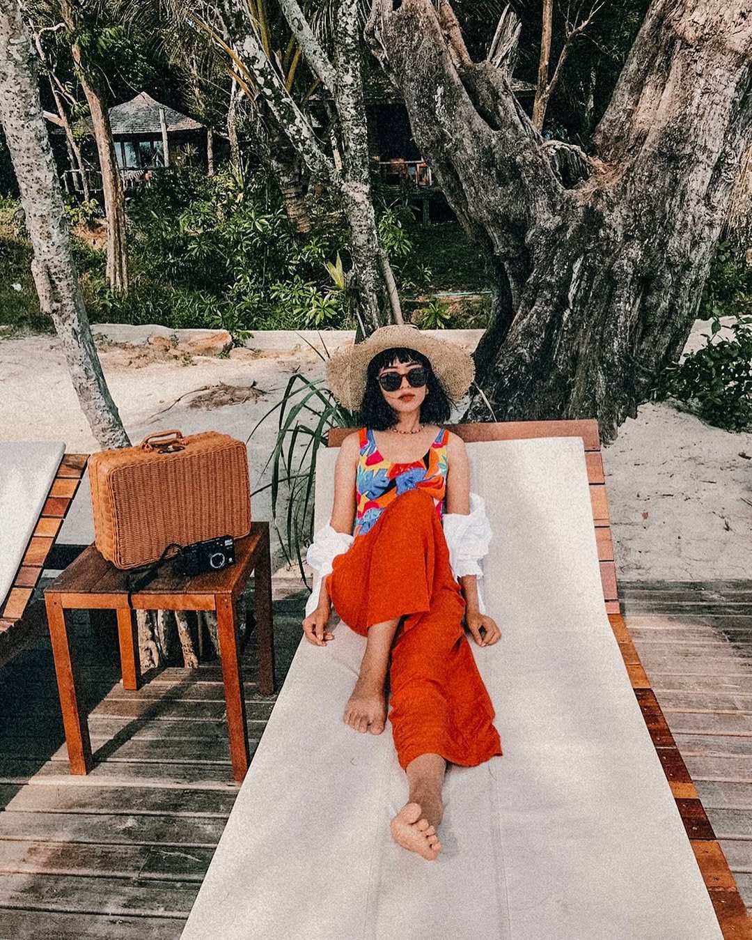Woman in a colorful top and red thai wrap pants skirt straw hat sitting on a lounge chair with a suitcase next to her on a wooden deck.