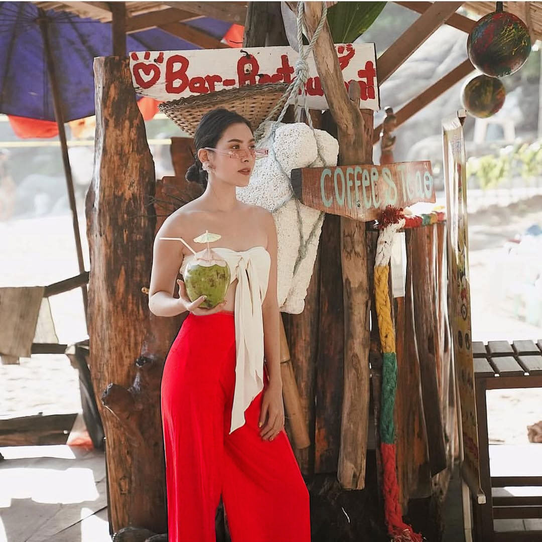 Woman in red Thai wrap pants skirt and white top holding a coconut in a rustic setting with wooden structures and signs.