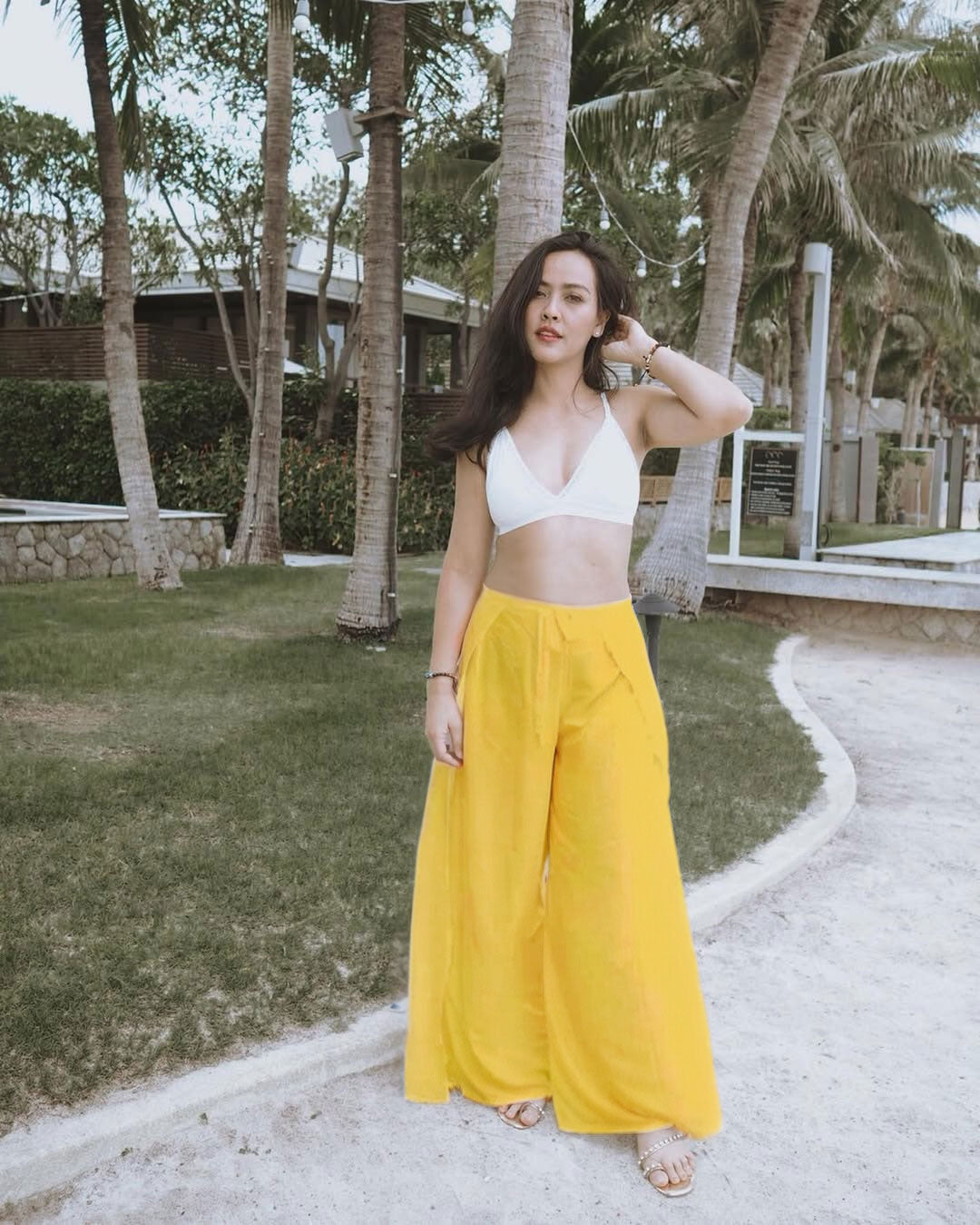 Woman in a white top and yellow thai wrap pants skirt standing on a path with palm trees in the background