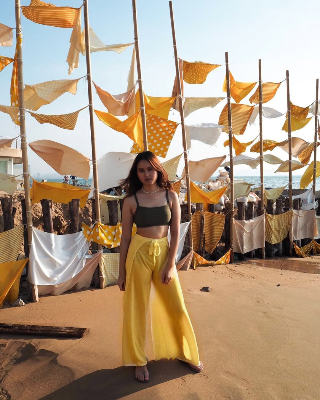 Woman in yellow thai wrap pants skirt standing in front of a decorative wall with hanging fabric and wooden sticks.