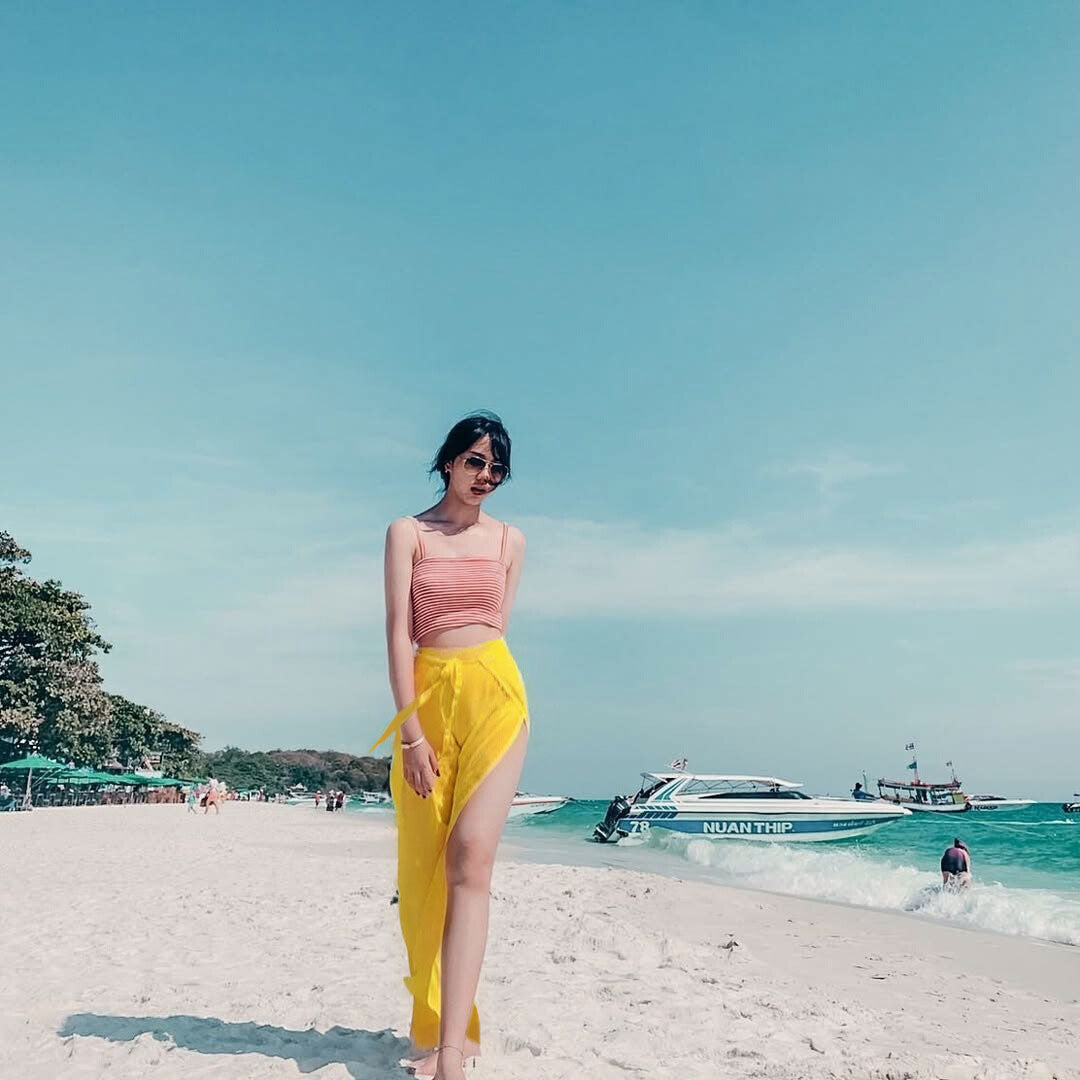 Woman in yellow thai wrap pants skirt and pink top standing on a beach with boats in the background
