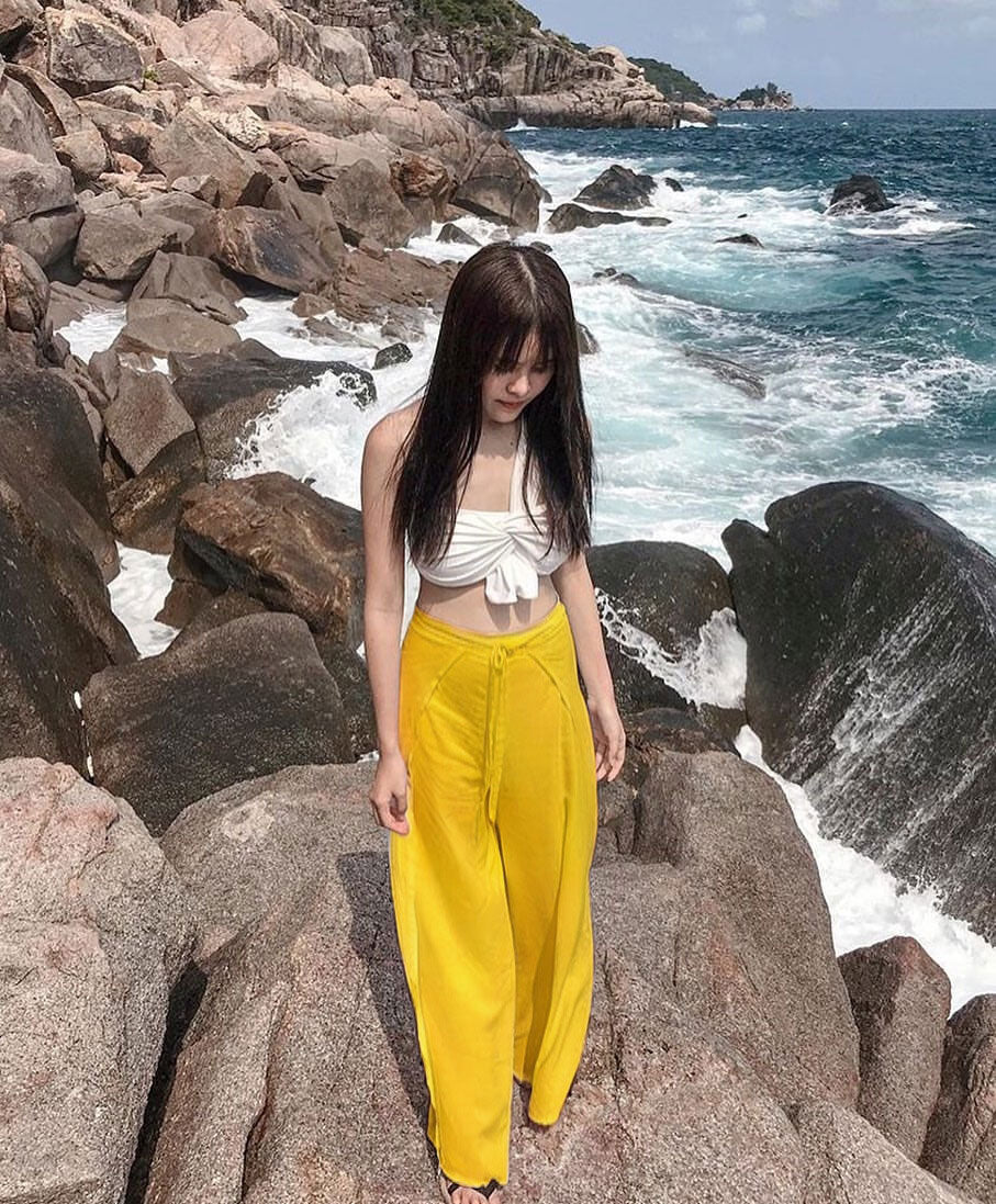 Woman in a white top and yellow thai wrap pants skirt standing on rocky coastal terrain with ocean waves.