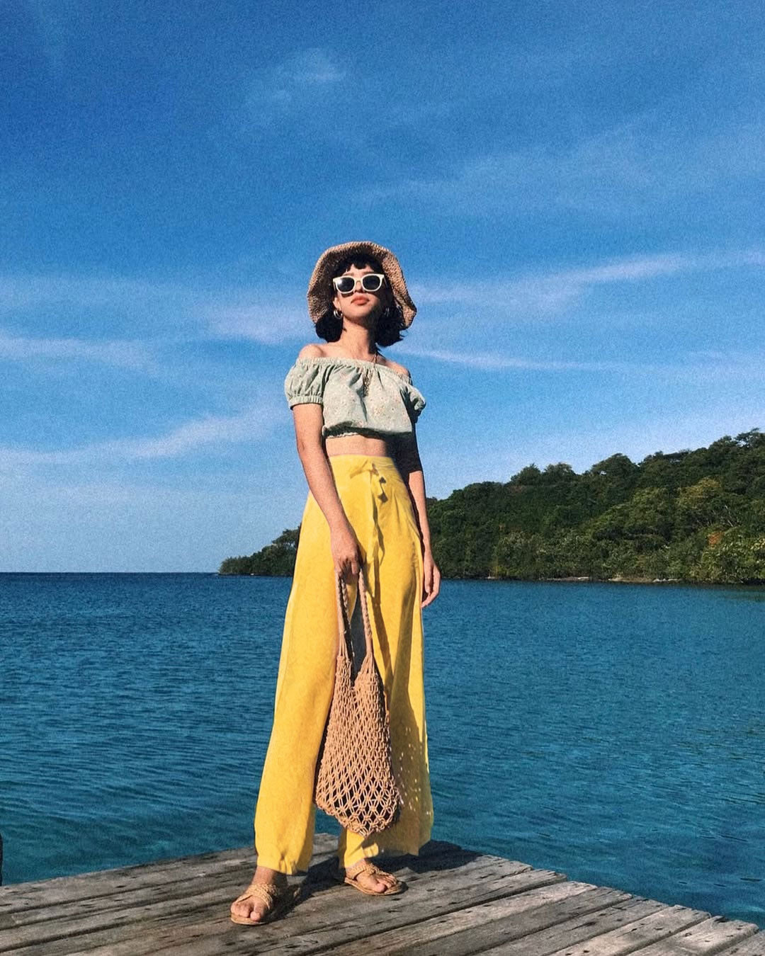 Woman in a green top and yellow thai wrap pants skirt standing on a dock with blue water and sky in the background