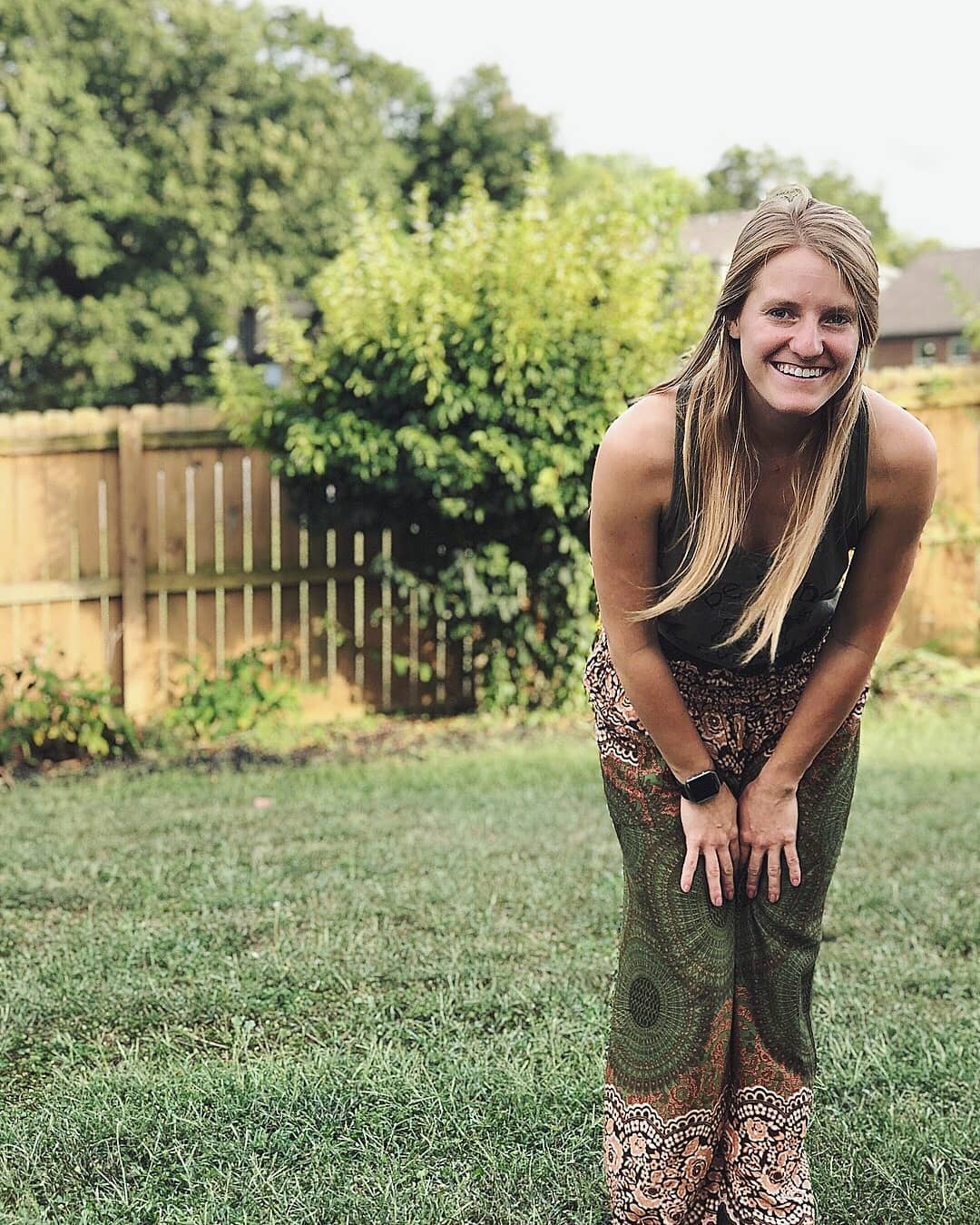 Woman wearing olive green Thai harem yoga pants with mandala print and green top posing outdoors in a garden with greenery in the background