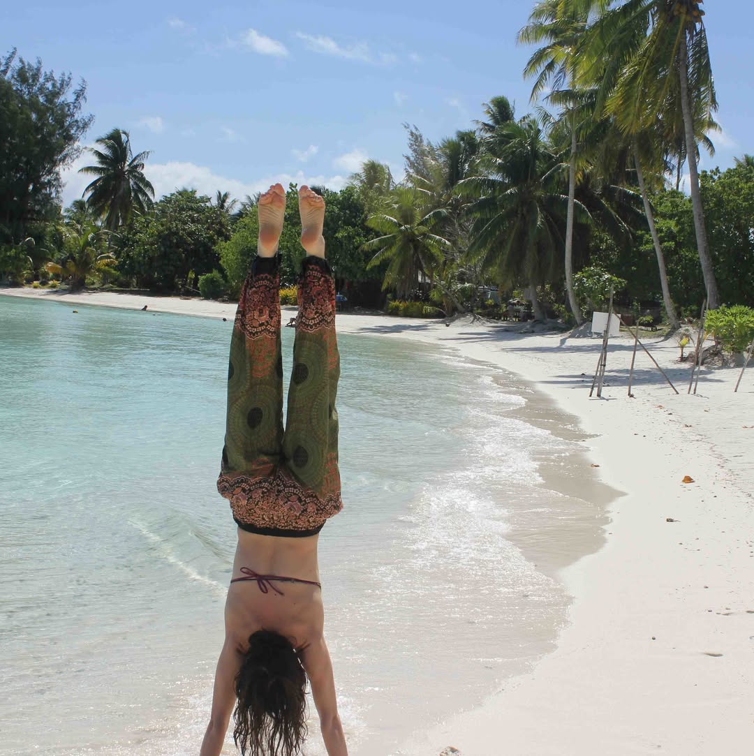 Woman wearing olive green Thai harem yoga pants with mandala print and bikini top doing a handstand on a tropical beach with palm trees and clear water.