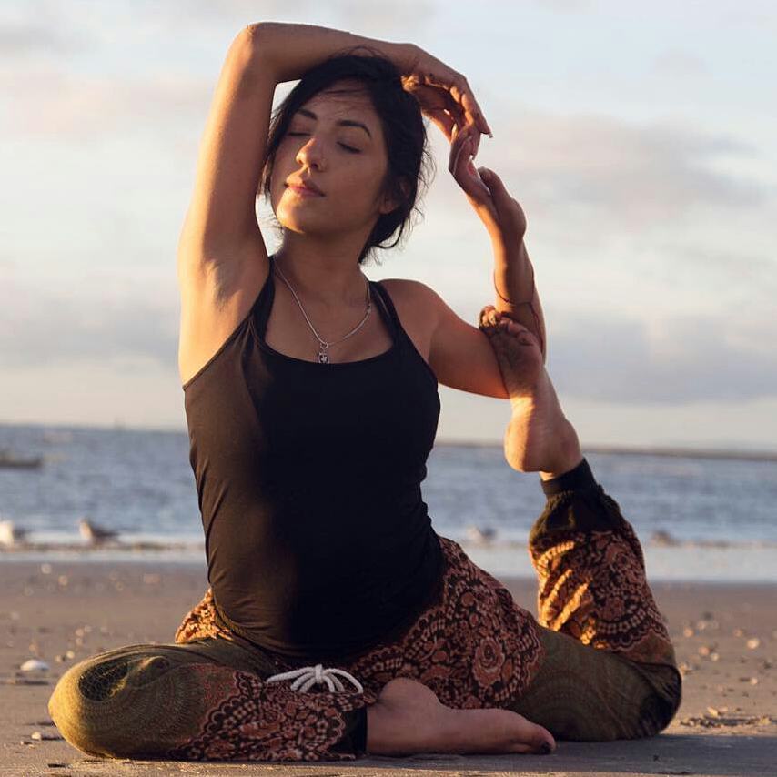 Woman wearing olive green Thai harem yoga pants with mandala print and black top practicing yoga on a beach at sunset