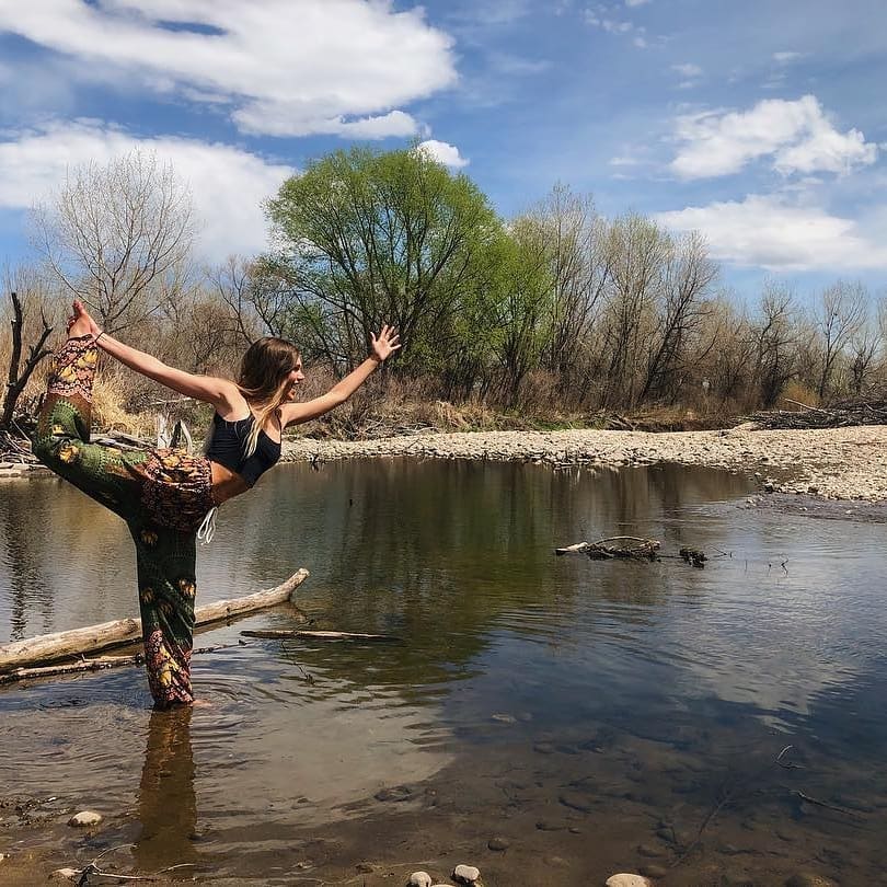 Woman wearing olive green Thai harem yoga pants with mandala print and black top practicing yoga by a pond with trees and blue sky in the background