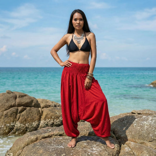 Woman in black bikini top and red thai harem pants standing on rocks by the ocean.