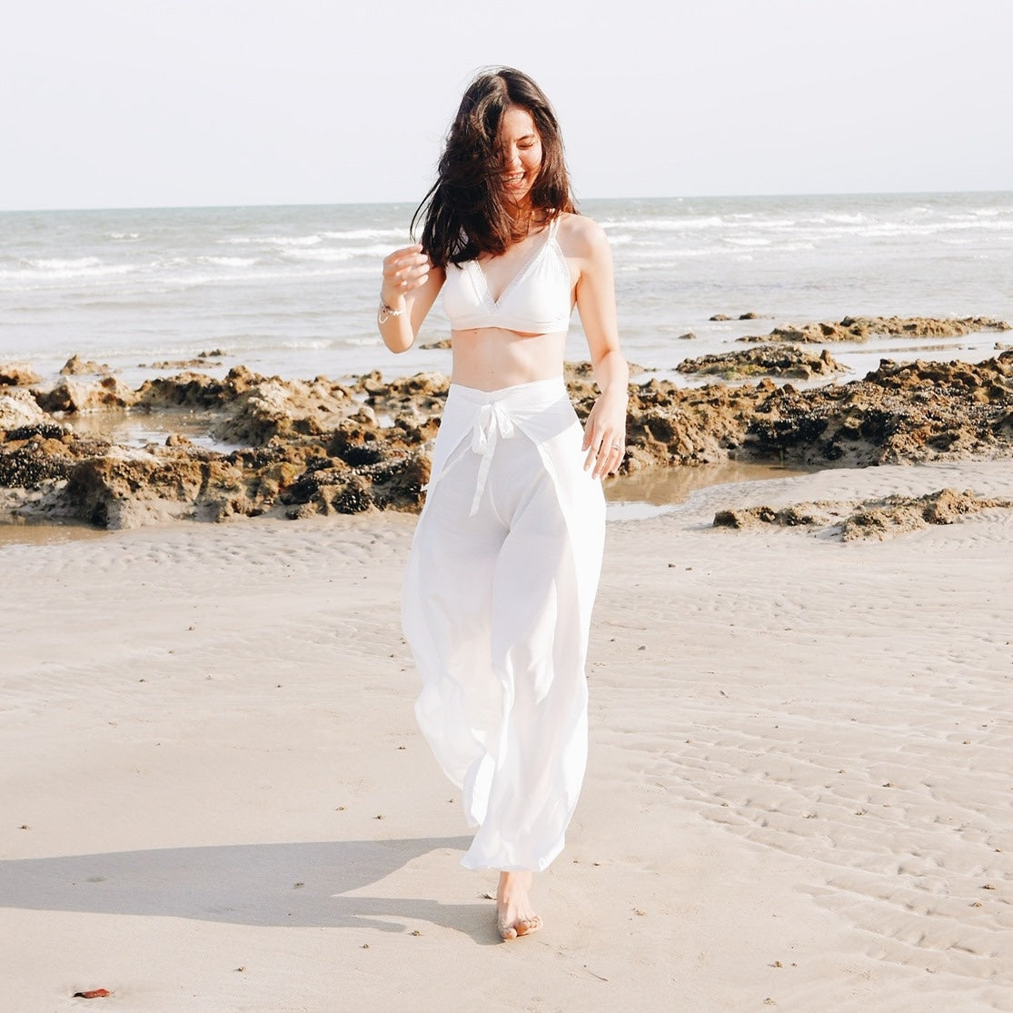 Woman in white thai wrap pants skirt and white top standing on a beach with rocks in the background