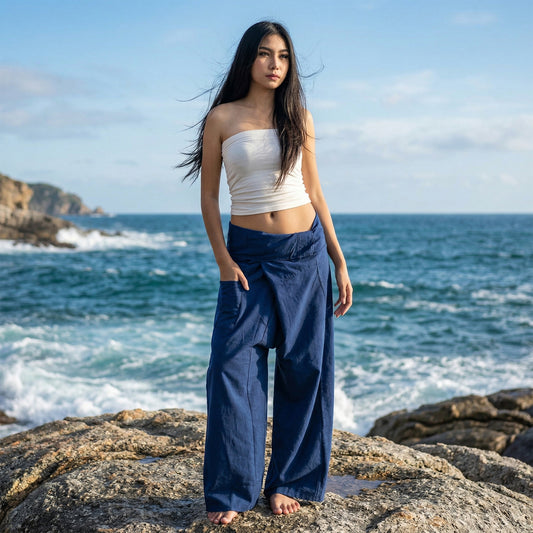 Female model standing confidently on rocks by the ocean, wearing versatile Moonlight Blue women's Thai fisherman pants.