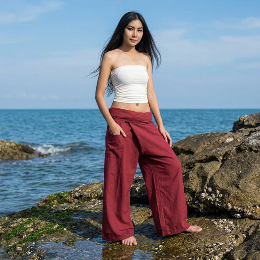 Female model standing confidently on rocks by the ocean, wearing versatile Mystic Maroon women's Thai fisherman pants.