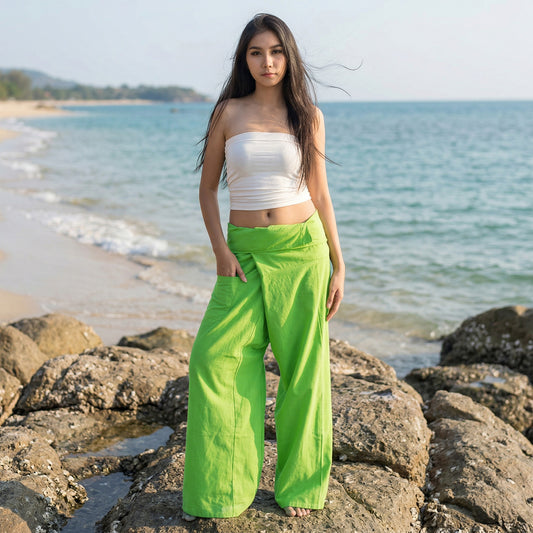 Female model standing confidently on rocks by the ocean, wearing versatile Tropical Lime women's Thai fisherman pants.