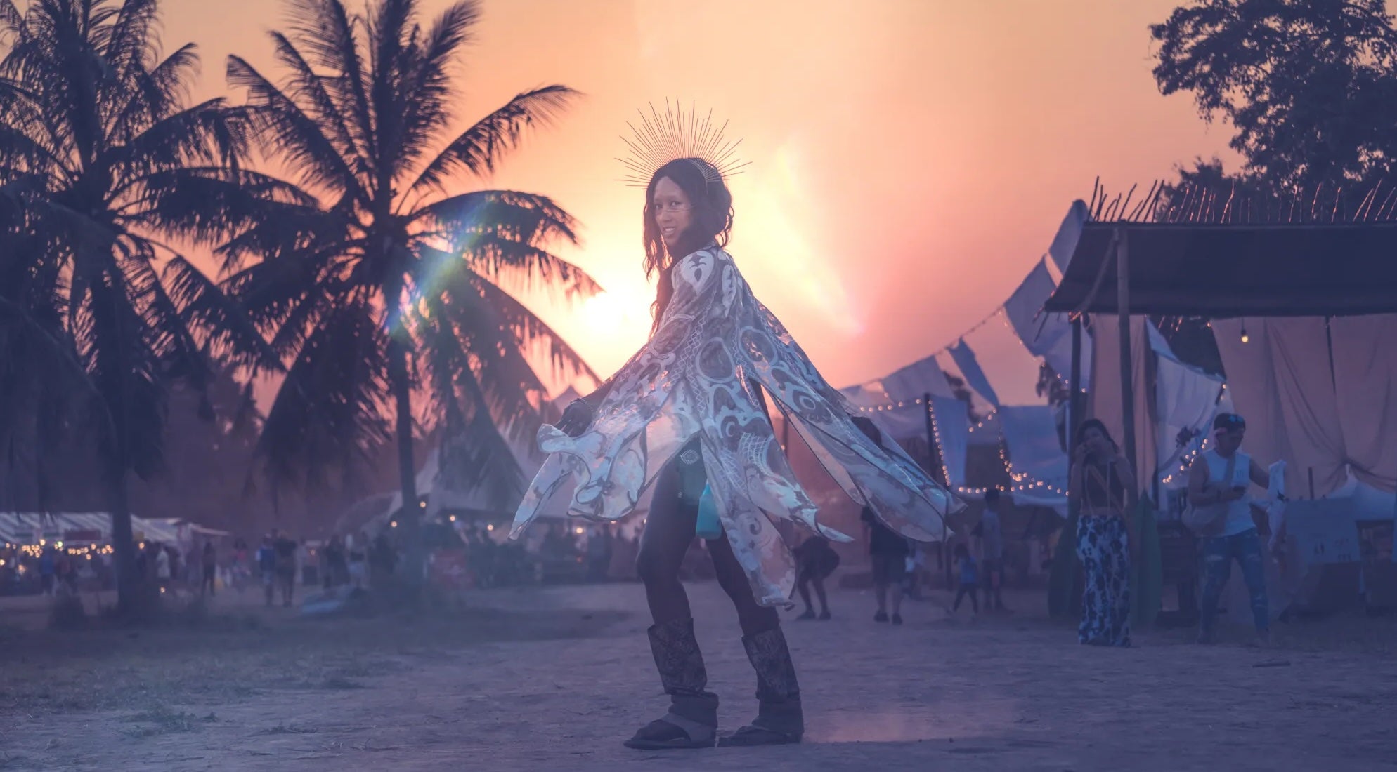 Person in a white outfit standing in a sandy area with palm trees and a sunset on Tomorrowland Thailand Festival