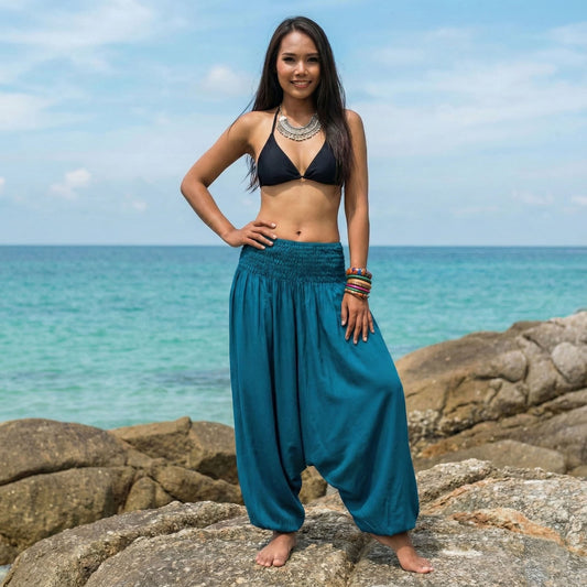 Woman in turquoise thai harem pants jumpsuit and black bikini top standing on rocks by the ocean