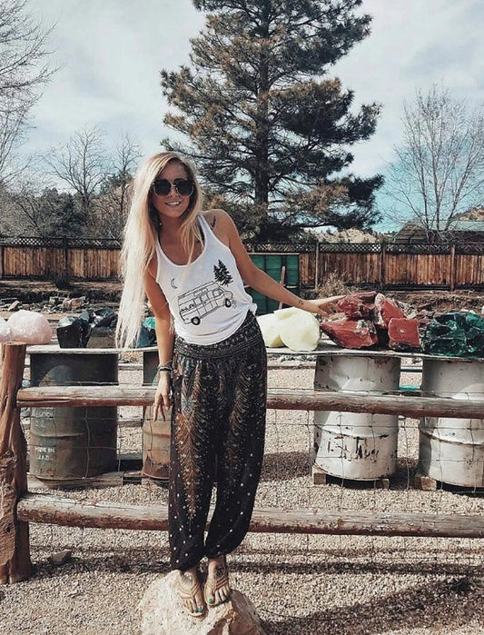 Woman standing outdoors near a wooden fence with various items in the background wearing green Thai harem yoga pants with peacock print and white top