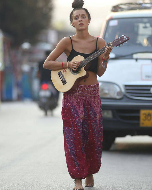 Woman walking on a street holding a guitar wearing a purple Thai harem yoga pants with peacock print and black top