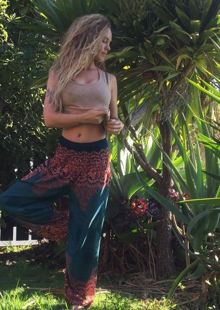 Woman in a flowing outfit standing in a lush green garden wearing turquoise Thai harem yoga pants with mandala print and grey top